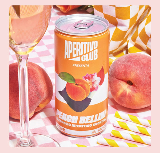 canette de pêche bellini posée sur une table avec nappe à carreaux, verre à pied rempli de cocktail, deux pêches et pailles jaunes