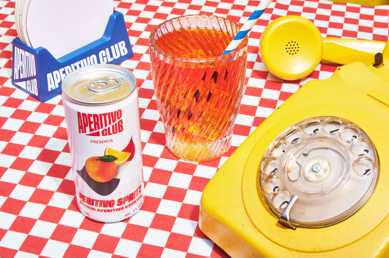 canette de spritz servie dans un verre rainuré avec paille, sur une table avec nappe à carreaux rouges, un téléphone jaune vintage à cadran rotatif