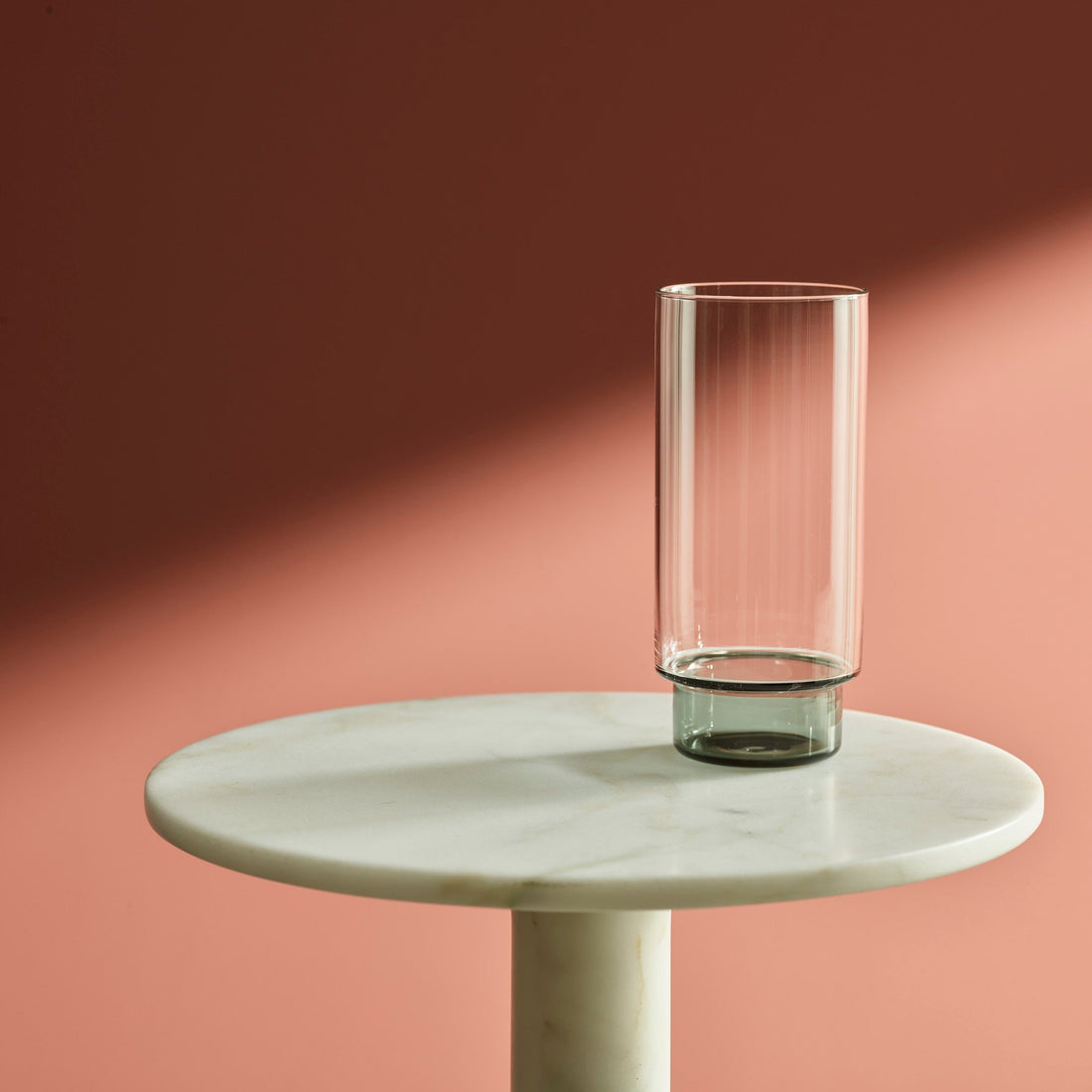 verre gris transparent vide posé sur une table en marbre blanc, mur de couleur vieux rose