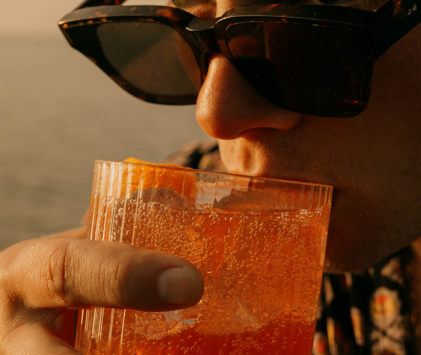 Homme portant des lunettes de soleil dégustant un mocktail orange sanguine dans un verre avec glaçons et agrume, lors de la golden hour au coucher du soleil