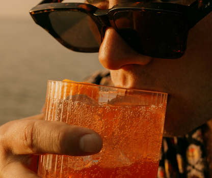 Homme portant des lunettes de soleil dégustant un mocktail orange sanguine dans un verre avec glaçons et agrume, lors de la golden hour au coucher du soleil